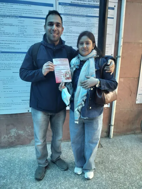 Couple holding a marriage-registration leaflet beside public information boards